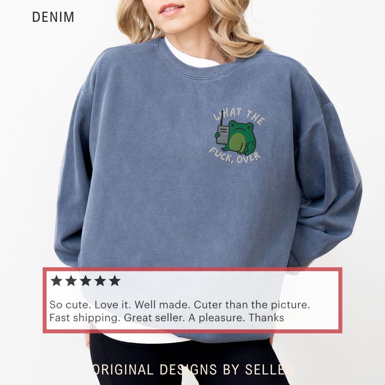 May include: Denim blue sweatshirt with the text "WHAT THE FUCK, OVER" and a cartoon frog holding a coffee cup. The sweatshirt has a crew neck and long sleeves. The word "DENIM" is printed at the top left.
