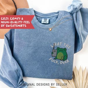 May include: A blue sweatshirt with the phrase "WHAT THE FUCK, OVER" and a green frog graphic. The sweatshirt also has the text "COZY, COMFY & HIGH-QUALITY FEEL OF SWEATSHIRTS" and "ORIGINAL DESIGNS BY SELLER". A gold necklace is draped on the sweatshirt.