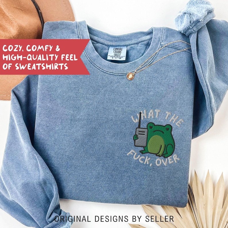 May include: A blue sweatshirt with the phrase "WHAT THE FUCK, OVER" and a green frog graphic. The sweatshirt also has the text "COZY, COMFY & HIGH-QUALITY FEEL OF SWEATSHIRTS" and "ORIGINAL DESIGNS BY SELLER". A gold necklace is draped on the sweatshirt.