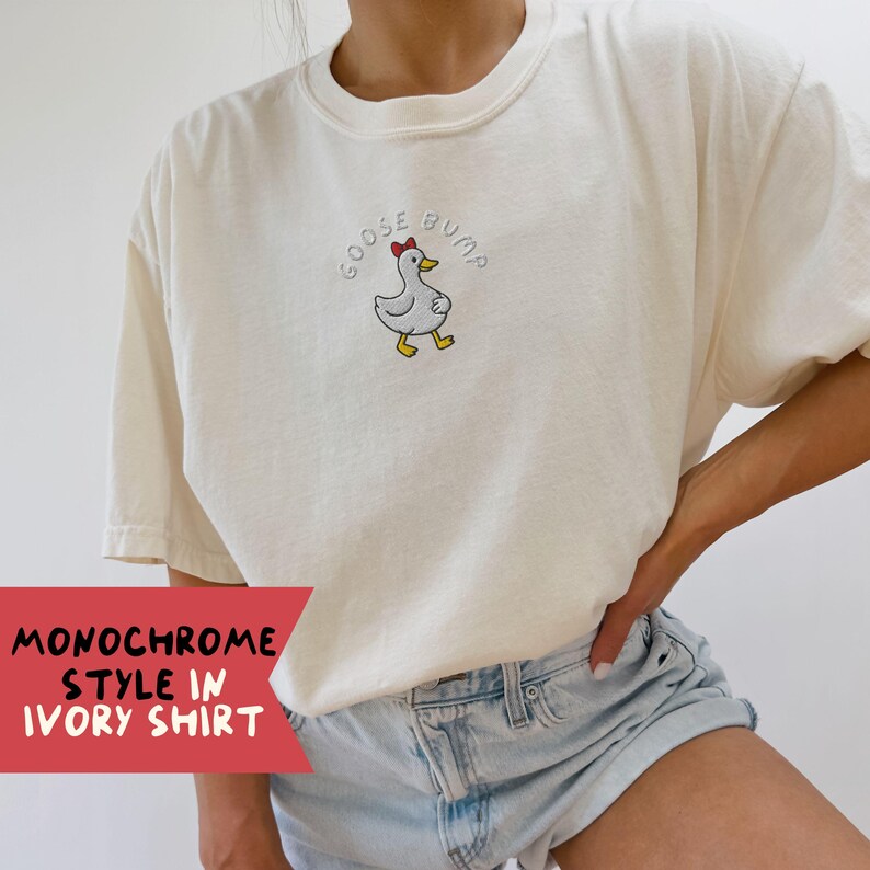 May include: Ivory-colored t-shirt with the embroidered phrase "GOOSE BUMP" above a cartoon duck wearing a red bow. The shirt is part of a monochrome style collection. The text "MONOCHROME STYLE IN IVORY SHIRT" is printed on a red banner.