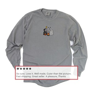 May include: Gray long-sleeve shirt with a small embroidered Halloween design featuring a ghost, a grim reaper, and a campfire. The shirt has a crew neck and a relaxed fit. The shirt is made of a soft, comfortable material.