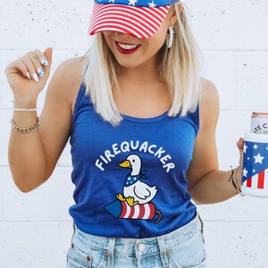 May include: A blue tank top with the text "FIREQUACKER" and a cartoon duck riding a firecracker. The duck wears a bandana. The person is wearing a red, white, and blue baseball cap and holding a can with a patriotic design.