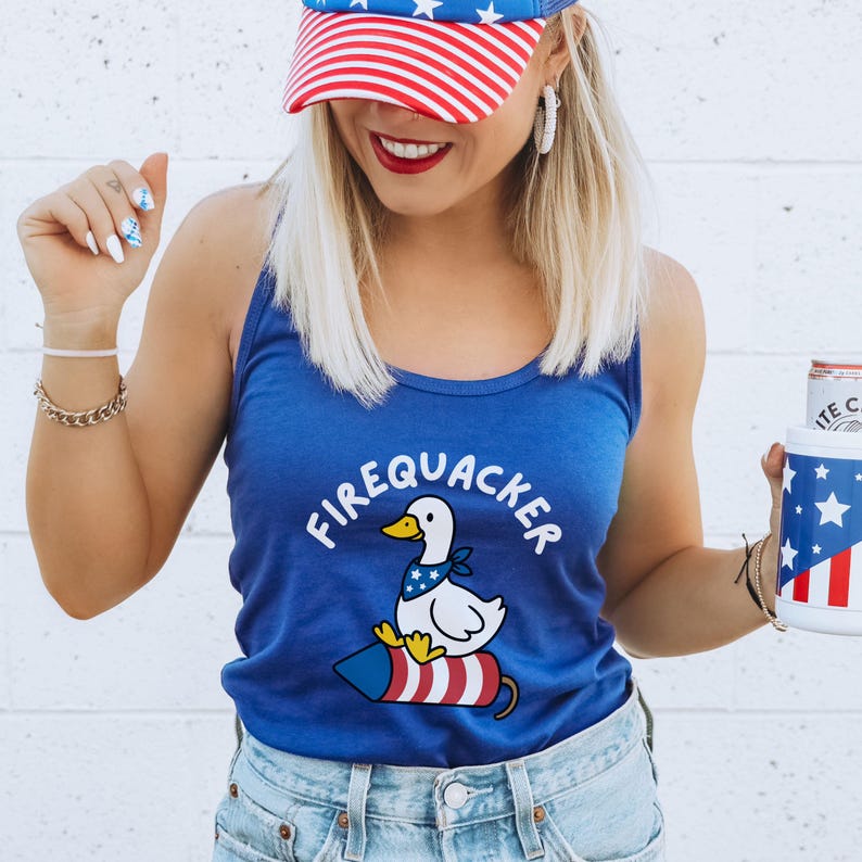May include: A blue tank top with the text "FIREQUACKER" and a cartoon duck riding a firecracker. The duck wears a bandana. The person is wearing a red, white, and blue baseball cap and holding a can with a patriotic design.