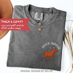 May include: A gray Comfort Colors t-shirt with the embroidered phrase "Silly Moose" and a cartoon moose. The shirt has the text "THICK & COMFY nice weight while still super comfy" and "ORIGINAL DESIGNS BY SELLER". A gold necklace is draped on the shirt.
