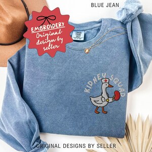 May include: Blue jean-colored sweatshirt with the text "KIDNEY SQUAD" and an embroidered goose wearing a nurse's hat and holding a kidney. The sweatshirt has the text "EMBROIDERY Original design by seller".