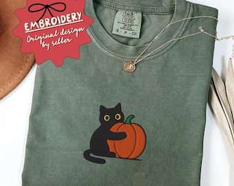 Pode incluir: Camiseta verde oliva com bordado de um gato preto abraçando uma abóbora laranja. A camiseta tem uma etiqueta Comfort Colors e um colar de prata. Uma etiqueta vermelha com "BORDADO" está no canto superior esquerdo.