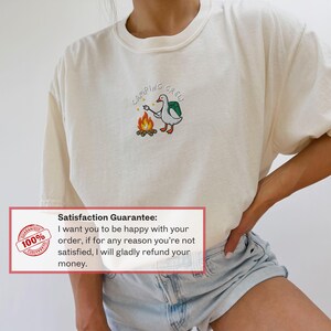 Op de afbeelding: Cr&egrave;mekleurig T-shirt met een geborduurd ontwerp van een gans die een marshmallow roostert boven een kampvuur, met de tekst "CAMPING CREW". Het shirt is gemaakt van een zacht materiaal.