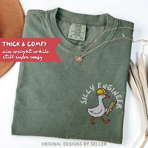 May include: An olive green t-shirt with the words "Silly Engineer" and a cartoon goose wearing a yellow hard hat and carrying blueprints. The shirt also has the text "Thick & Comfy" and "nice weight while still super comfy". A gold necklace is draped across the shirt.