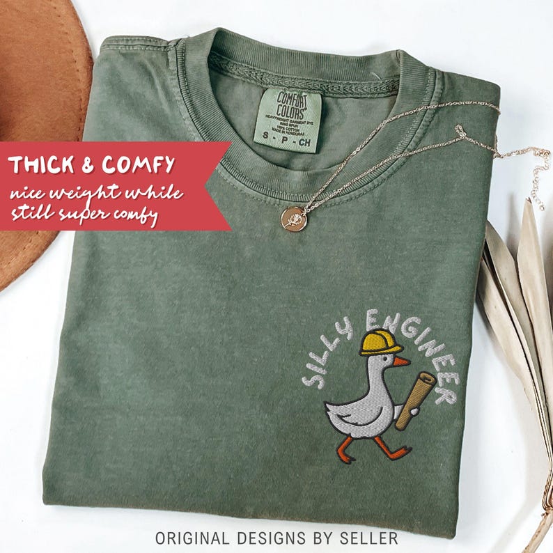 May include: An olive green t-shirt with the words "Silly Engineer" and a cartoon goose wearing a yellow hard hat and carrying blueprints. The shirt also has the text "Thick & Comfy" and "nice weight while still super comfy". A gold necklace is draped across the shirt.