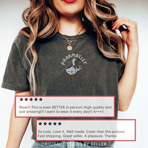 May include: Dark gray t-shirt featuring an embroidered design of a goose wearing a pharmacist's coat and holding a medicine bottle. The word "PHARMACIST" is arched above the goose. The shirt has customer reviews.