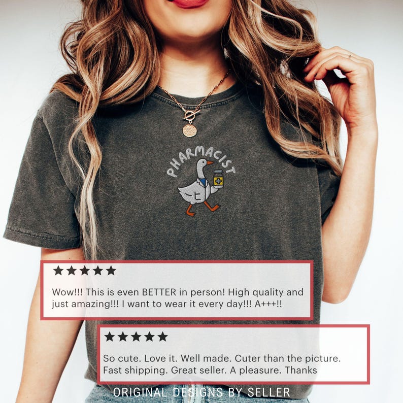 May include: Dark gray t-shirt featuring an embroidered design of a goose wearing a pharmacist's coat and holding a medicine bottle. The word "PHARMACIST" is arched above the goose. The shirt has customer reviews.