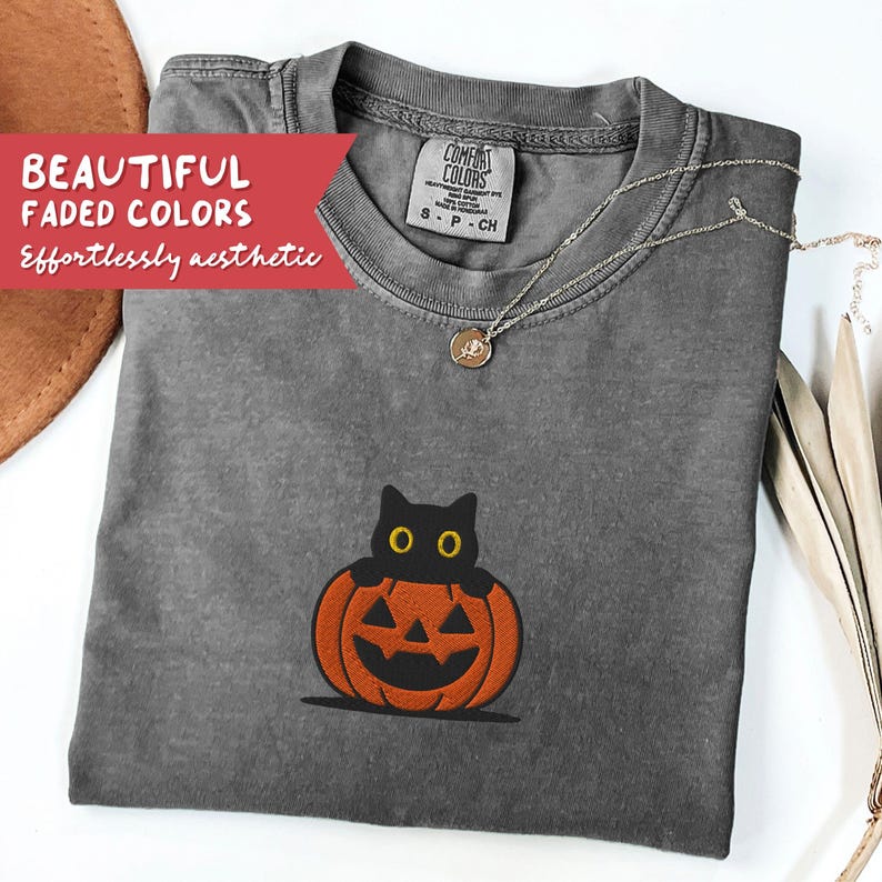 K&ouml;nnte beinhalten: Ein graues Comfort Colors T-Shirt mit einer schwarzen Katze, die &uuml;ber einen orangefarbenen K&uuml;rbis schaut. Das Shirt hat einen Rundhalsausschnitt und kurze &Auml;rmel. Eine silberne Halskette mit Anh&auml;nger ist auf dem Shirt drapiert. Der Text "BEAUTIFUL FADED COLORS" und "Effortlessly aesthetic" befindet sich auf einem roten Banner.