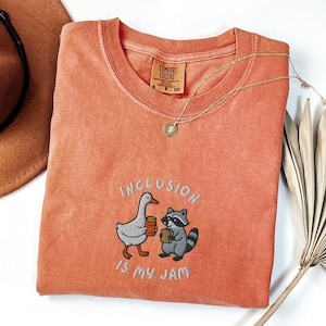 K&ouml;nnte beinhalten: Korallenfarbenes T-Shirt mit dem gestickten Schriftzug "INCLUSION IS MY JAM" und Illustrationen einer Gans und eines Waschb&auml;ren, die Marmelade halten. Eine goldene Halskette und ein brauner Hut sind ebenfalls zu sehen.