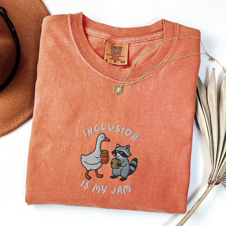 K&ouml;nnte beinhalten: Korallenfarbenes T-Shirt mit dem gestickten Schriftzug "INCLUSION IS MY JAM" und Illustrationen einer Gans und eines Waschb&auml;ren, die Marmelade halten. Eine goldene Halskette und ein brauner Hut sind ebenfalls zu sehen.