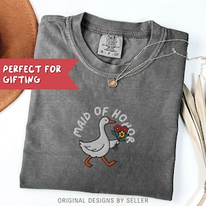 May include: A gray Comfort Colors t-shirt with the embroidered words "MAID OF HONOR" and a cartoon goose carrying flowers. The shirt has a crew neck and short sleeves. A gold necklace with a pendant is draped on the shirt. The text "ORIGINAL DESIGNS BY SELLER" is at the bottom.