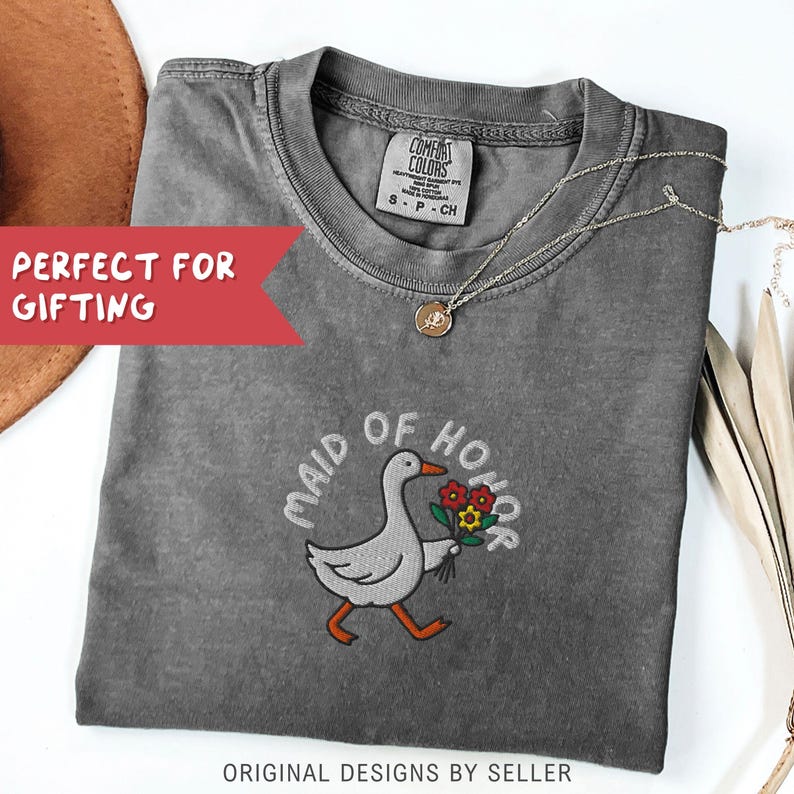 May include: A gray Comfort Colors t-shirt with the embroidered words "MAID OF HONOR" and a cartoon goose carrying flowers. The shirt has a crew neck and short sleeves. A gold necklace with a pendant is draped on the shirt. The text "ORIGINAL DESIGNS BY SELLER" is at the bottom.