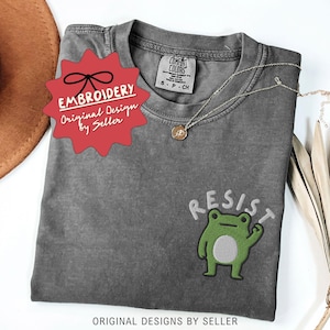 May include: A gray t-shirt with an embroidered green frog design and the word "RESIST" in white. The shirt has a small tag that says "COMFORT COLORS". A red tag with the word "EMBROIDERY" is also visible. The text "ORIGINAL DESIGNS BY SELLER" is at the bottom.