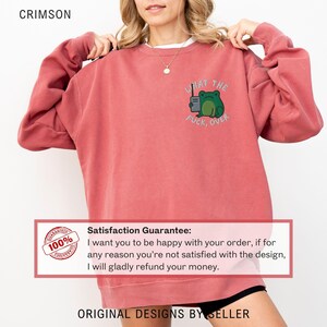 May include: Crimson-colored sweatshirt with a frog graphic holding a radio and the text "WHAT THE FUCK, OVER." The sweatshirt is a relaxed fit and has long sleeves. The text "ORIGINAL DESIGNS BY SELLER" is at the bottom.