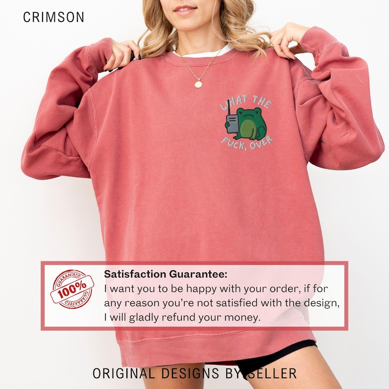 May include: Crimson-colored sweatshirt with a frog graphic holding a radio and the text "WHAT THE FUCK, OVER." The sweatshirt is a relaxed fit and has long sleeves. The text "ORIGINAL DESIGNS BY SELLER" is at the bottom.