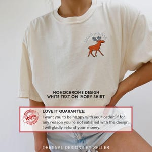 May include: Ivory-colored t-shirt featuring a cartoon moose with the text "Silly Moose." The shirt has a monochrome design with white text. The shirt is made of a soft material.