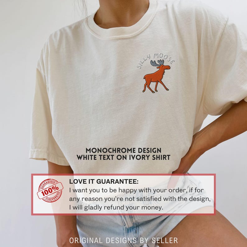 May include: Ivory-colored t-shirt featuring a cartoon moose with the text "Silly Moose." The shirt has a monochrome design with white text. The shirt is made of a soft material.