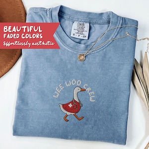 May include: A light blue Comfort Colors t-shirt with the embroidered phrase "WEE WOO CREW" and a cartoon goose wearing a red medical vest and stethoscope. A gold necklace with a pendant is draped on the shirt. The text "BEAUTIFUL FADED COLORS" is on a red banner.