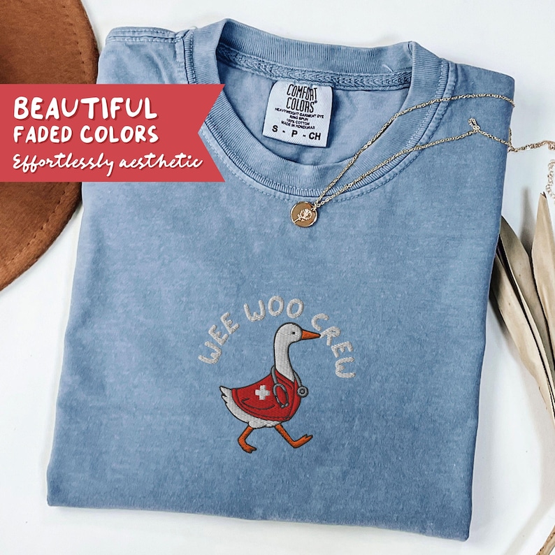May include: A light blue Comfort Colors t-shirt with the embroidered phrase "WEE WOO CREW" and a cartoon goose wearing a red medical vest and stethoscope. A gold necklace with a pendant is draped on the shirt. The text "BEAUTIFUL FADED COLORS" is on a red banner.