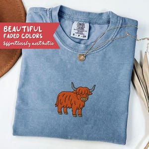 May include: A light blue Comfort Colors t-shirt with an embroidered orange Highland cow design. The shirt has a crew neck and short sleeves. A gold necklace with a pendant is draped on the shirt. Text on the shirt reads "BEAUTIFUL FADED COLORS Effortlessly aesthetic."