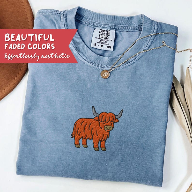 May include: A light blue Comfort Colors t-shirt with an embroidered orange Highland cow design. The shirt has a crew neck and short sleeves. A gold necklace with a pendant is draped on the shirt. Text on the shirt reads "BEAUTIFUL FADED COLORS Effortlessly aesthetic."