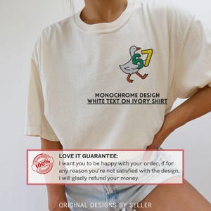 May include: Ivory-colored t-shirt featuring a cartoon duck carrying the number 67. The design includes the text "MONOCHROME DESIGN WHITE TEXT ON IVORY SHIRT". The shirt has a crew neck and short sleeves.