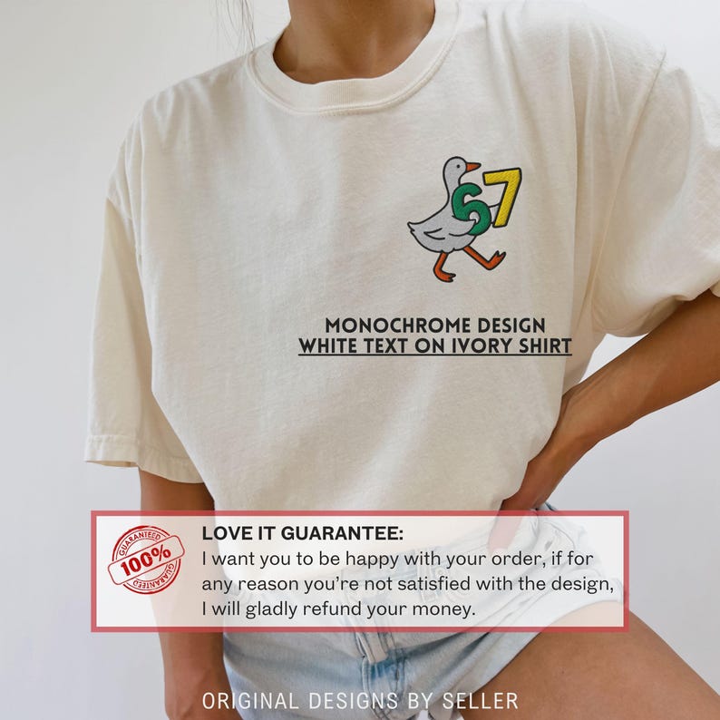 May include: Ivory-colored t-shirt featuring a cartoon duck carrying the number 67. The design includes the text "MONOCHROME DESIGN WHITE TEXT ON IVORY SHIRT". The shirt has a crew neck and short sleeves.
