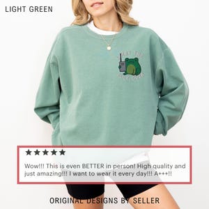 May include: Light green sweatshirt with a frog graphic holding a walkie-talkie and the text "WHAT THE FUCK, OVER." The sweatshirt is a light green color and has a crew neck. The text "ORIGINAL DESIGNS BY SELLER" is at the bottom.
