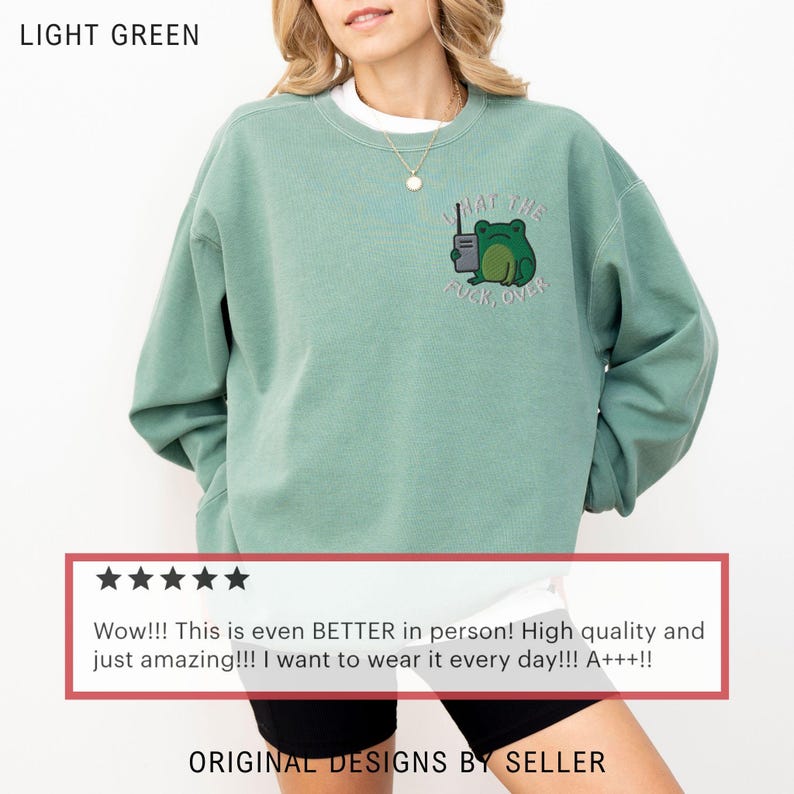 May include: Light green sweatshirt with a frog graphic holding a walkie-talkie and the text "WHAT THE FUCK, OVER." The sweatshirt is a light green color and has a crew neck. The text "ORIGINAL DESIGNS BY SELLER" is at the bottom.