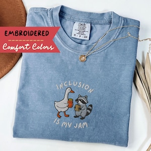 K&ouml;nnte beinhalten: Hellblaues Comfort Colors T-Shirt mit dem gestickten Schriftzug "Inclusion is my Jam". Das Design zeigt eine Gans und einen Waschb&auml;ren, die Tassen halten. Eine goldene Halskette mit Anh&auml;nger liegt auf dem Shirt.