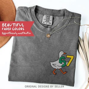 May include: A gray Comfort Colors t-shirt with a duck graphic holding the numbers 6 and 7. The shirt has the text "BEAUTIFUL FADED COLORS" and "Effortlessly aesthetic". A gold necklace is draped across the shirt. The text "ORIGINAL DESIGNS BY SELLER" is at the bottom.