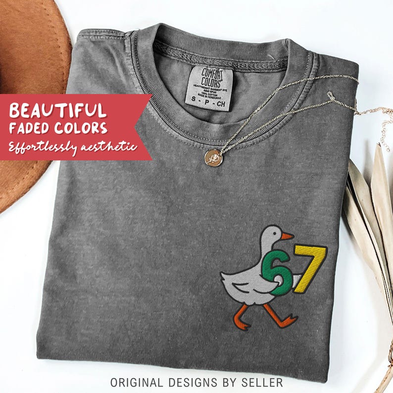 May include: A gray Comfort Colors t-shirt with a duck graphic holding the numbers 6 and 7. The shirt has the text "BEAUTIFUL FADED COLORS" and "Effortlessly aesthetic". A gold necklace is draped across the shirt. The text "ORIGINAL DESIGNS BY SELLER" is at the bottom.
