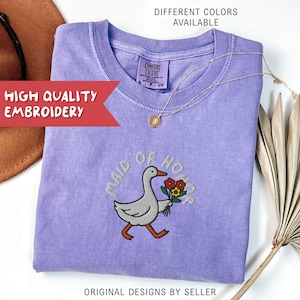 May include: A lilac-colored t-shirt with the embroidered phrase "MAID OF HONOR" and a goose carrying flowers. The shirt has a crew neck and short sleeves. A gold necklace with a pendant is draped on the shirt. The text "DIFFERENT COLORS AVAILABLE" is at the top.
