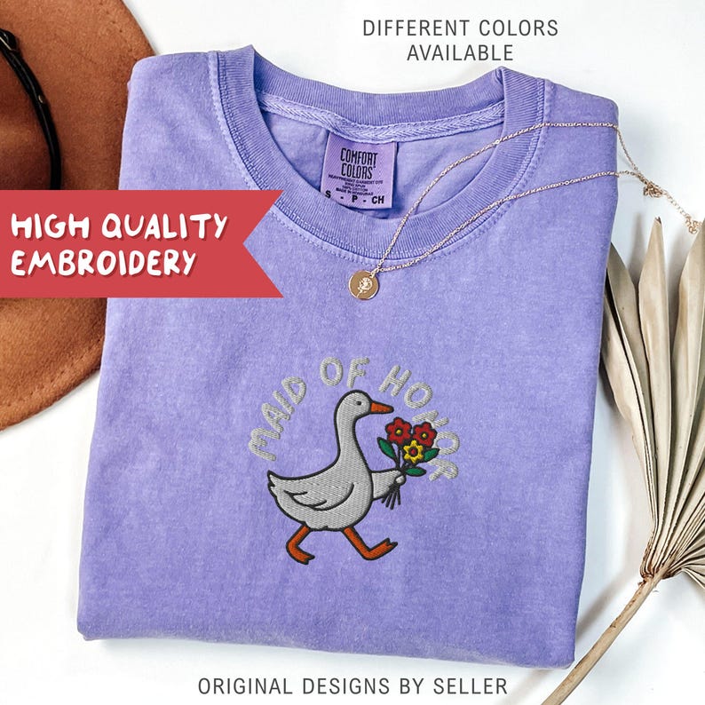 May include: A lilac-colored t-shirt with the embroidered phrase "MAID OF HONOR" and a goose carrying flowers. The shirt has a crew neck and short sleeves. A gold necklace with a pendant is draped on the shirt. The text "DIFFERENT COLORS AVAILABLE" is at the top.