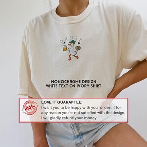 May include: Ivory-colored t-shirt with a cartoon goose design holding a beer and pretzel, wearing a hat. The word "PROST" is above the goose. Text on the shirt reads "MONOCHROME DESIGN WHITE TEXT ON IVORY SHIRT".