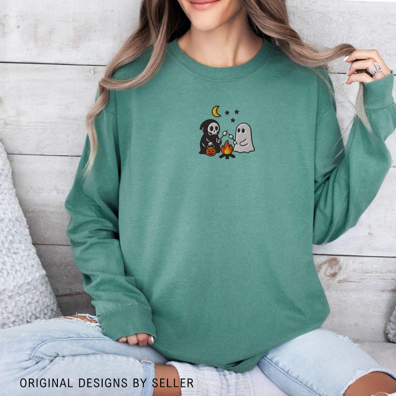 May include: Teal green sweatshirt with a Halloween-themed embroidered design. The design features a black Grim Reaper, a white ghost, a campfire, a crescent moon, and stars. The text "ORIGINAL DESIGNS BY SELLER" is at the bottom.