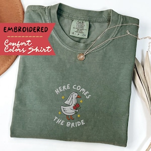 May include: An olive green Comfort Colors shirt with embroidered text and a graphic of a duck wearing a veil and holding roses. The text reads "HERE COMES THE BRIDE". A gold necklace is draped on the shirt.
