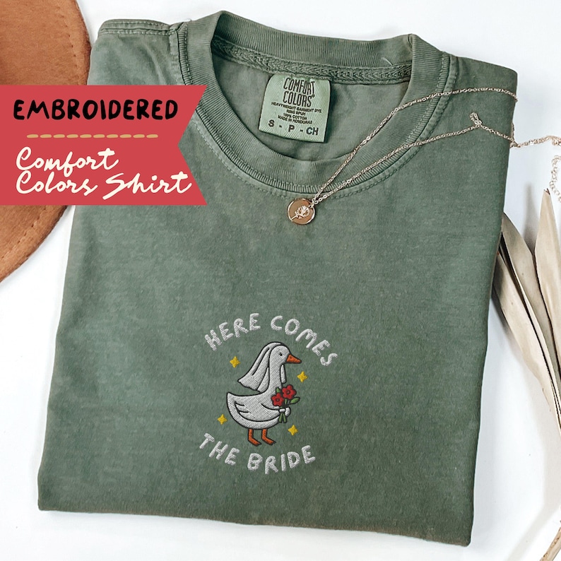 May include: An olive green Comfort Colors shirt with embroidered text and a graphic of a duck wearing a veil and holding roses. The text reads "HERE COMES THE BRIDE". A gold necklace is draped on the shirt.