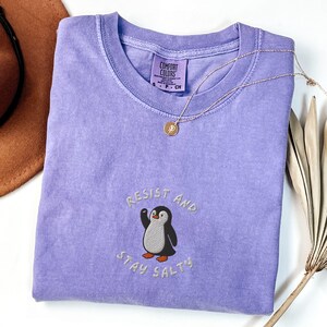Puede incluir: Una camiseta Comfort Colors de color lila claro con la frase bordada "Resist and Stay Salty" y un dise&ntilde;o de ping&uuml;ino. Un collar dorado con un colgante descansa sobre la camiseta. Un sombrero marr&oacute;n y una planta seca est&aacute;n en el fondo.