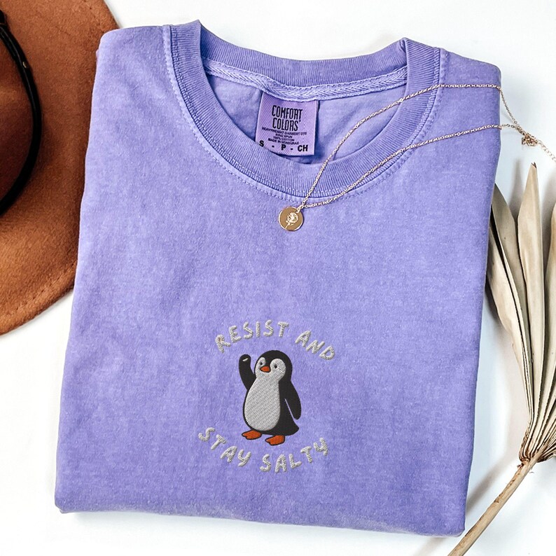 Puede incluir: Una camiseta Comfort Colors de color lila claro con la frase bordada "Resist and Stay Salty" y un dise&ntilde;o de ping&uuml;ino. Un collar dorado con un colgante descansa sobre la camiseta. Un sombrero marr&oacute;n y una planta seca est&aacute;n en el fondo.