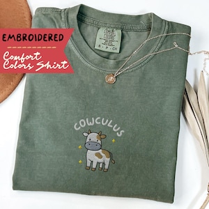 May include: An olive green Comfort Colors shirt with an embroidered design. The design features the word "COWCULUS" above a cartoon cow with brown and white spots. A gold necklace with a pendant rests on the shirt.