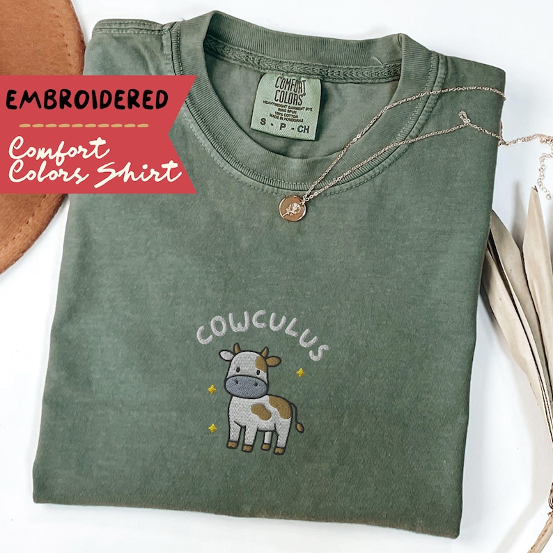 May include: An olive green Comfort Colors shirt with an embroidered design. The design features the word "COWCULUS" above a cartoon cow with brown and white spots. A gold necklace with a pendant rests on the shirt.