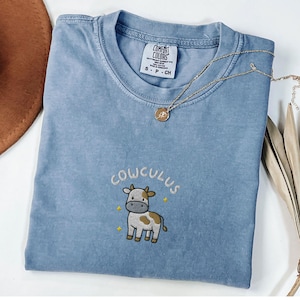 May include: A blue Comfort Colors t-shirt with the word "COWCULUS" embroidered above a cartoon cow. A gold necklace with a pendant rests on the shirt. The shirt has a crew neck and short sleeves.