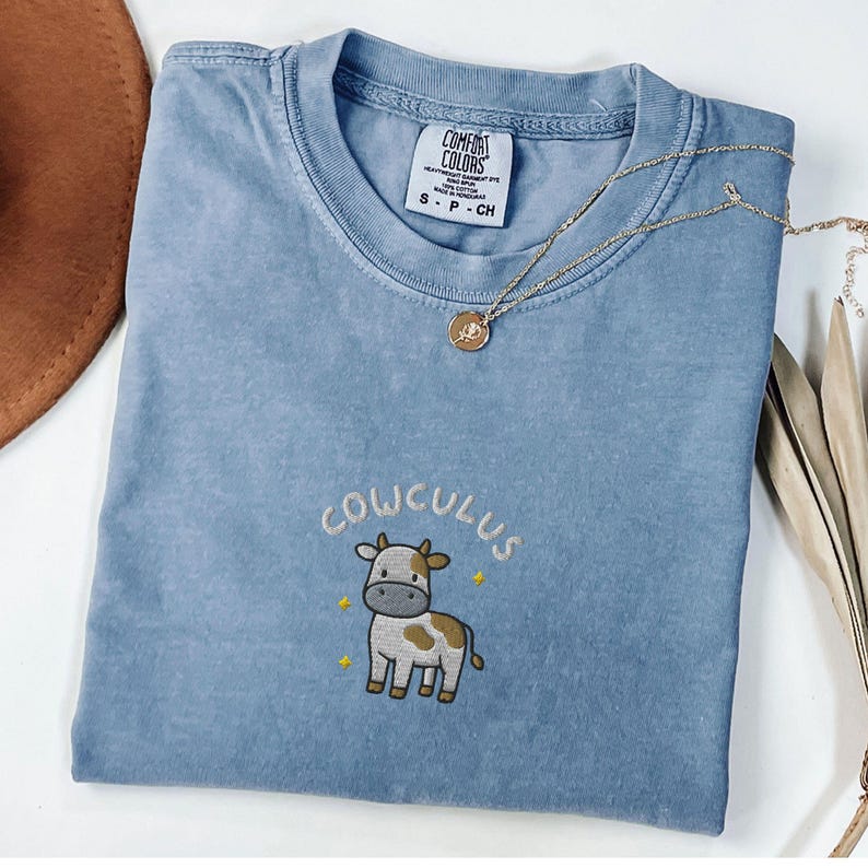 May include: A blue Comfort Colors t-shirt with the word "COWCULUS" embroidered above a cartoon cow. A gold necklace with a pendant rests on the shirt. The shirt has a crew neck and short sleeves.