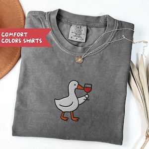 May include: A gray Comfort Colors t-shirt featuring an embroidered duck holding a glass of red wine. The shirt has a crew neck and short sleeves. A gold necklace with a pendant is draped on the shirt. The text "COMFORT COLORS SHIRTS" is on a red banner.