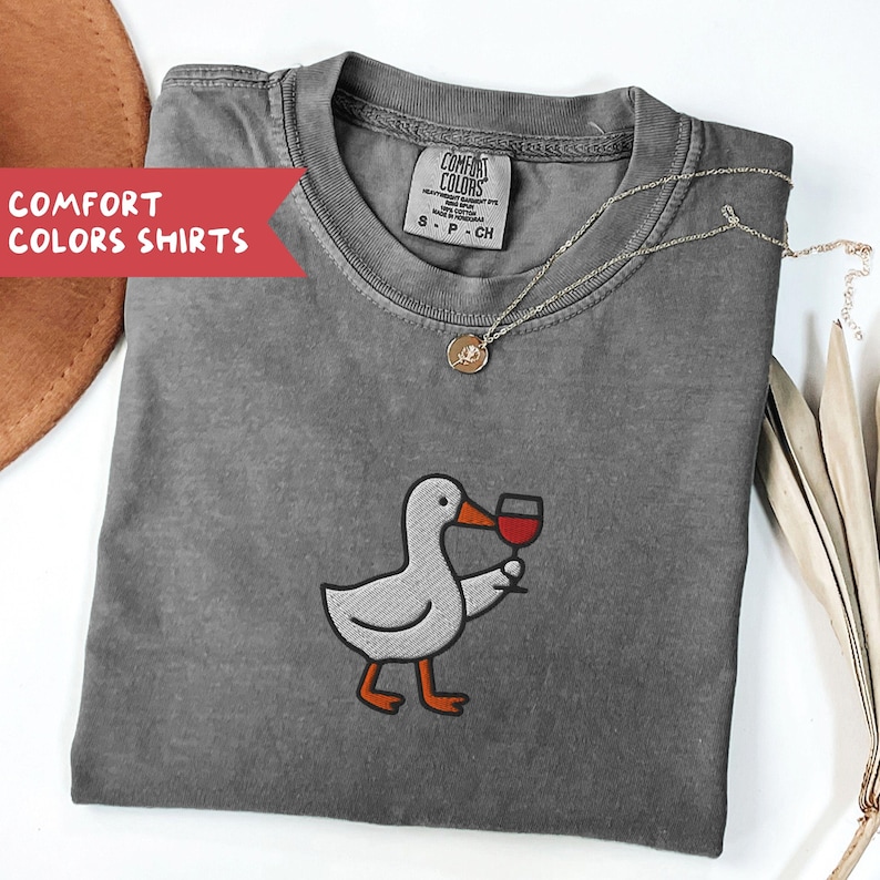 May include: A gray Comfort Colors t-shirt featuring an embroidered duck holding a glass of red wine. The shirt has a crew neck and short sleeves. A gold necklace with a pendant is draped on the shirt. The text "COMFORT COLORS SHIRTS" is on a red banner.